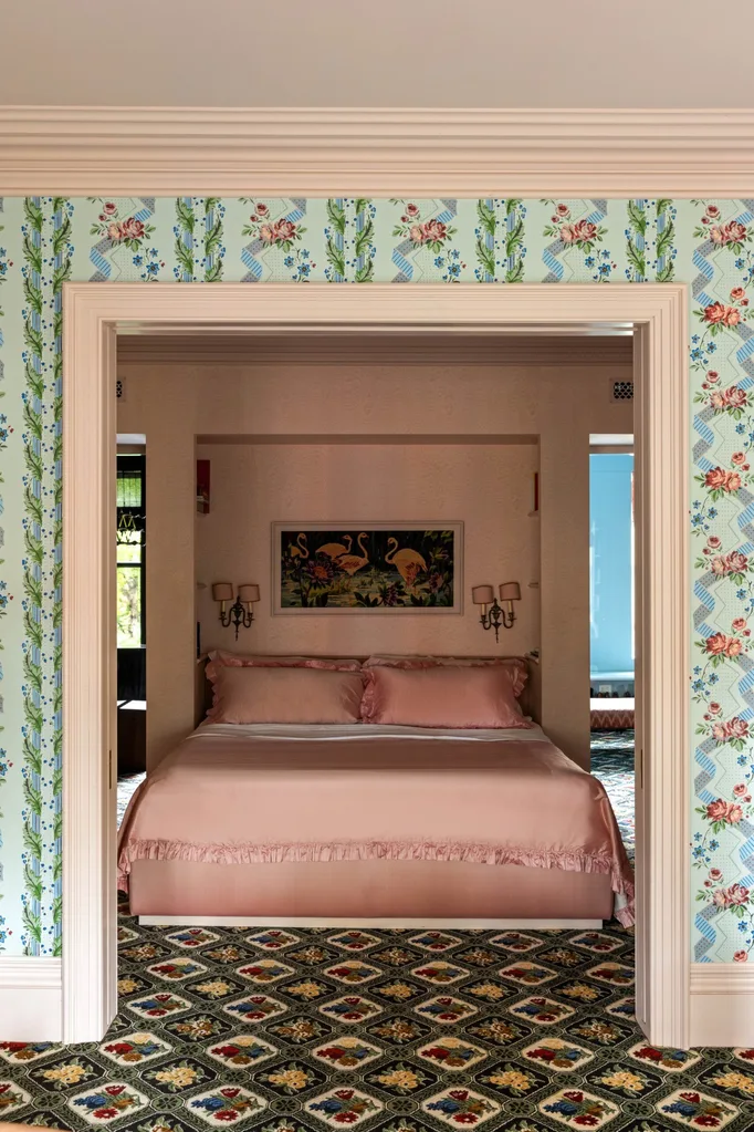 An open doorway, surrounded by floral wallpaper leads to a view of a bedroom with pink bedding, vintage wall art, and ornate patterned carpet.