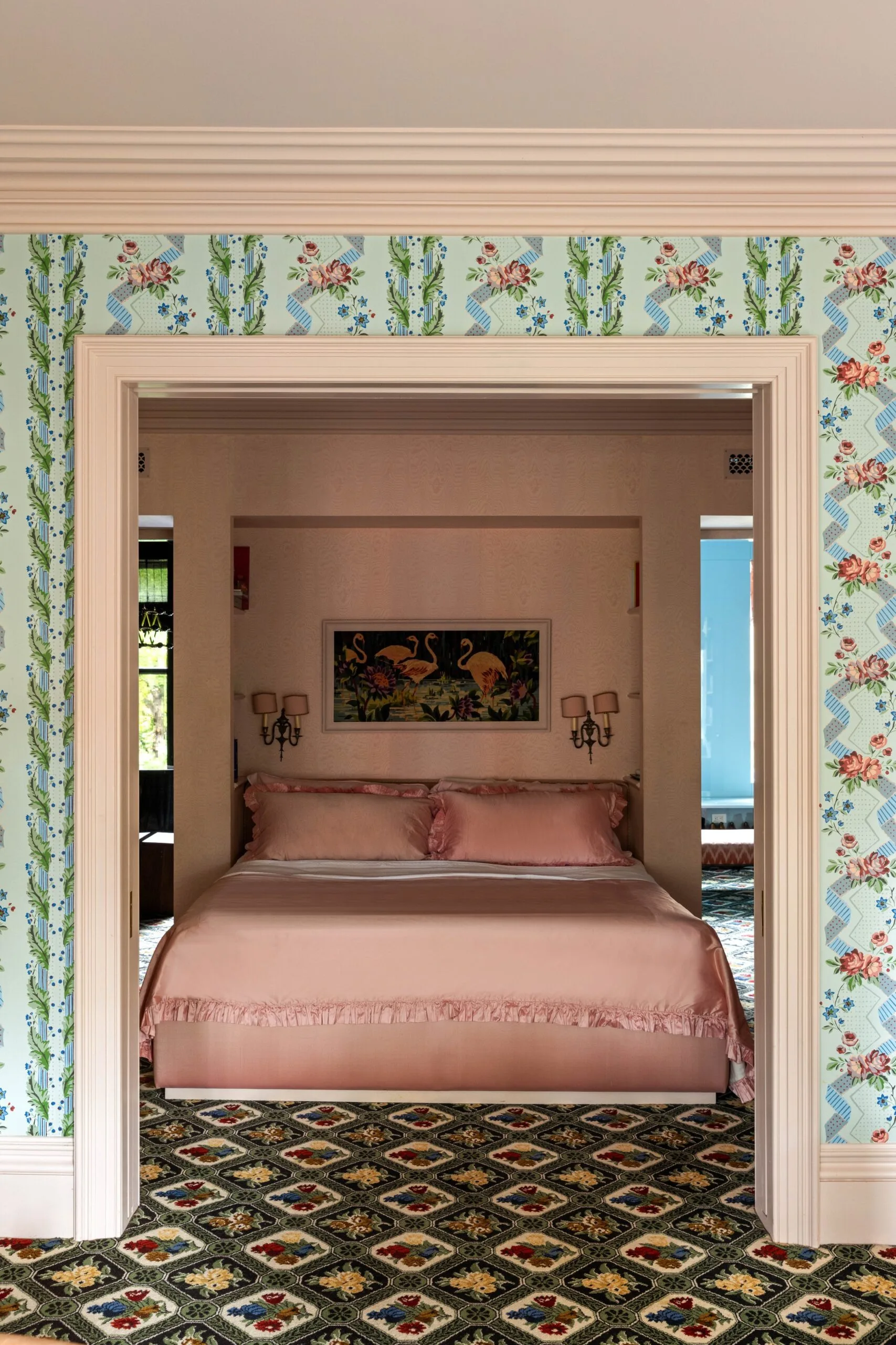 An open doorway, surrounded by floral wallpaper leads to a view of a bedroom with pink bedding, vintage wall art, and ornate patterned carpet. This is the bedroom of Lily Allen and David Harbour