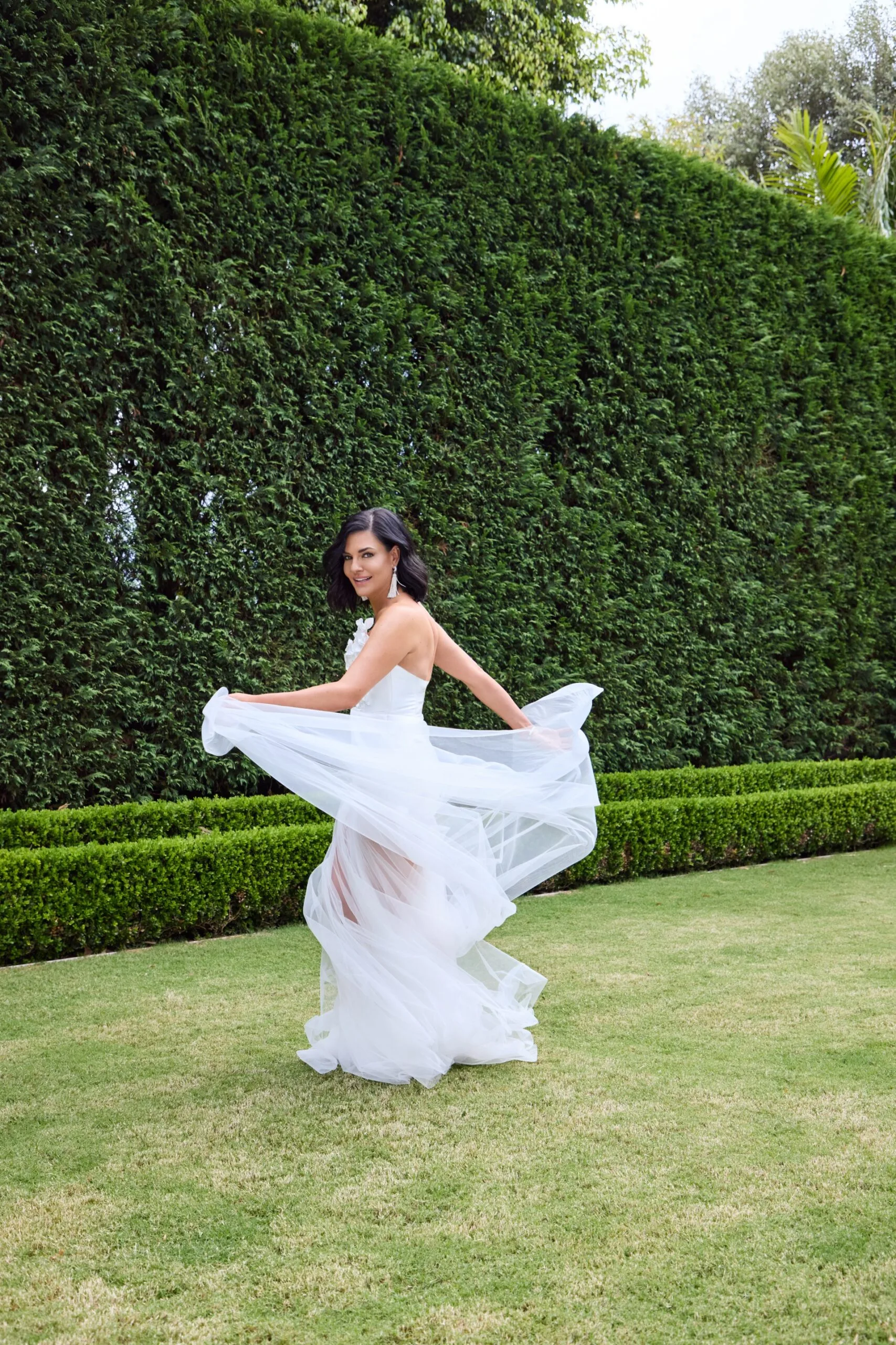 Nicole Gazal O'Neil dressed in a white formal gown in her backyard on the lawn with a hedge as a backdrop.