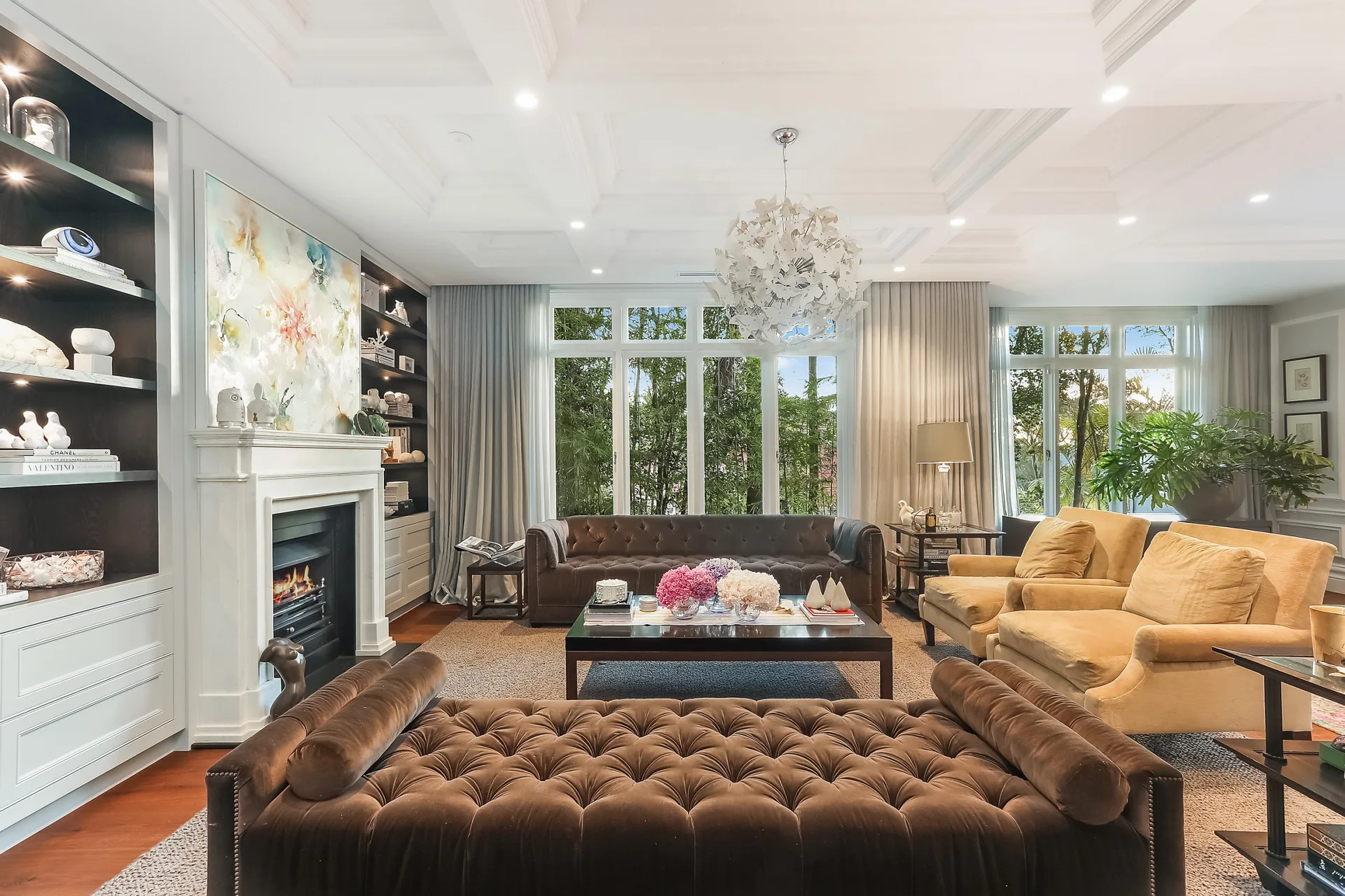 Nicole Gazal O'Neil's formal living room featuring a brown sofa, brown velvet daybed, camel-coloured armchairs, a butterfly chandelier, and windows with views to the garden.