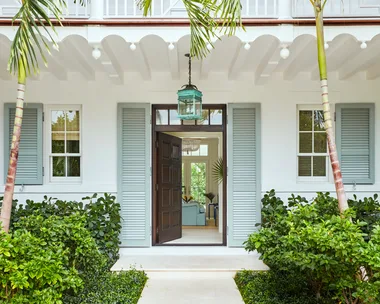 Entrance of a tropical-style house with open double doors, flanked by palm trees and lush greenery.