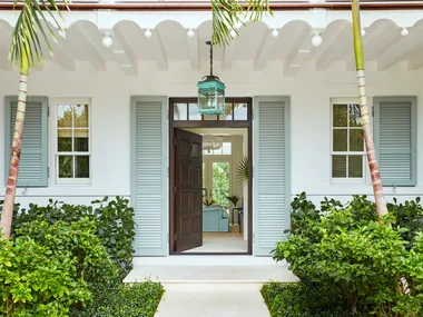 Entrance of a tropical-style house with open double doors, flanked by palm trees and lush greenery.