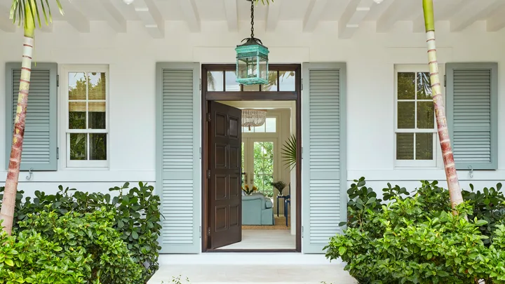 Entrance of a tropical-style house with open double doors, flanked by palm trees and lush greenery.