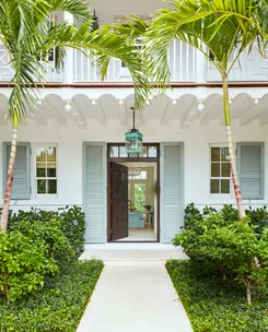 Entrance of a tropical-style house with open double doors, flanked by palm trees and lush greenery.