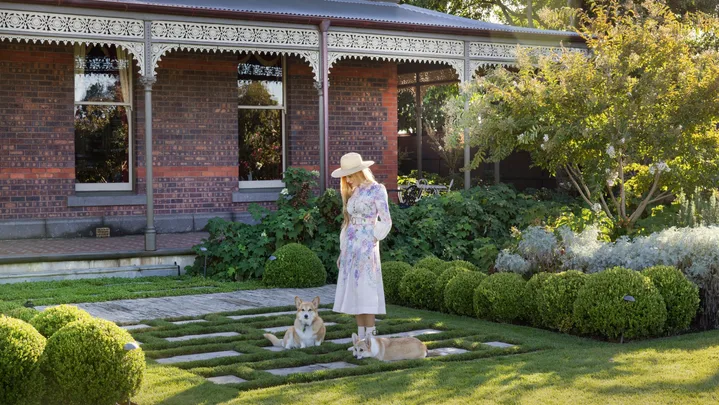 Woman in floral dress and hat standing in a garden with two corgis, brick house in the background. The garden was designed by Paul Bangay.