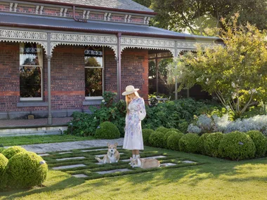 Woman in floral dress and hat standing in a garden with two corgis, brick house in the background. The garden was designed by Paul Bangay.