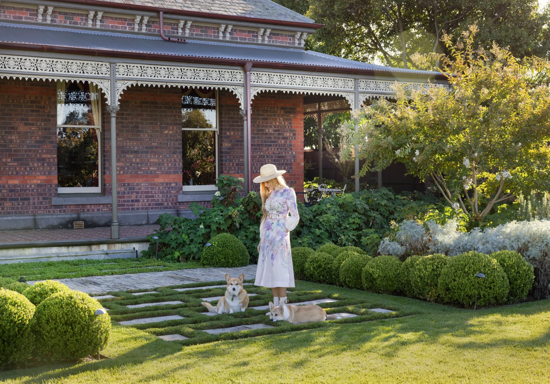 Woman in floral dress and hat standing in a garden with two corgis, brick house in the background. The garden was designed by Paul Bangay.