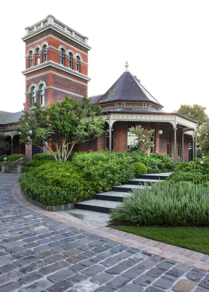 Victorian-style brick house with a tower, ornate trim, lush greenery, and cobblestone path in the foreground.