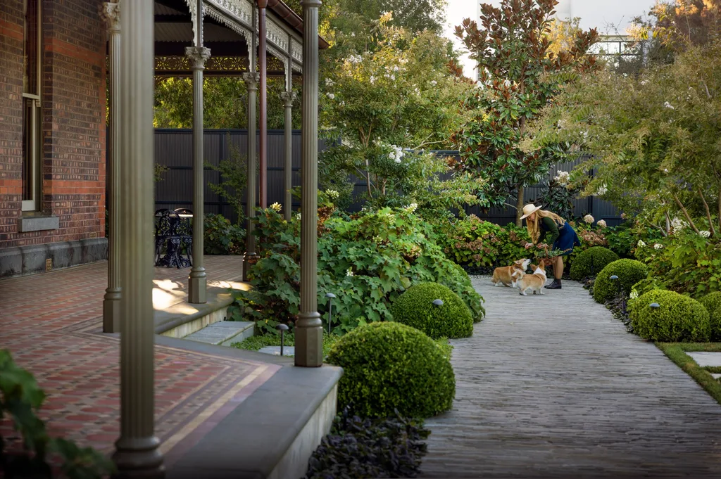 Victorian house veranda, lush garden, woman in hat petting two dogs, corgis, on a stone path.