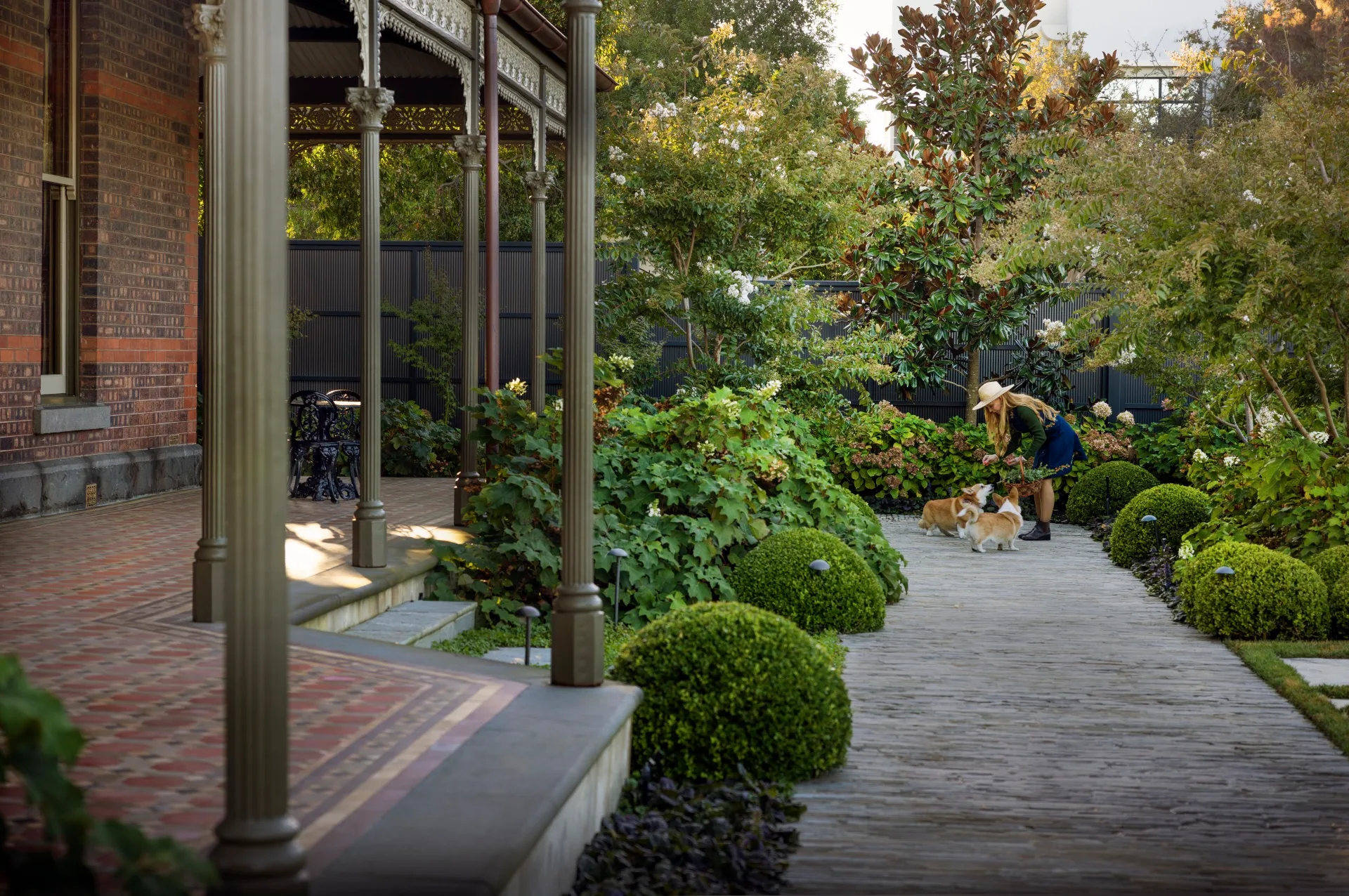 Victorian house veranda, lush garden, woman in hat petting two dogs, corgis, on a stone path.