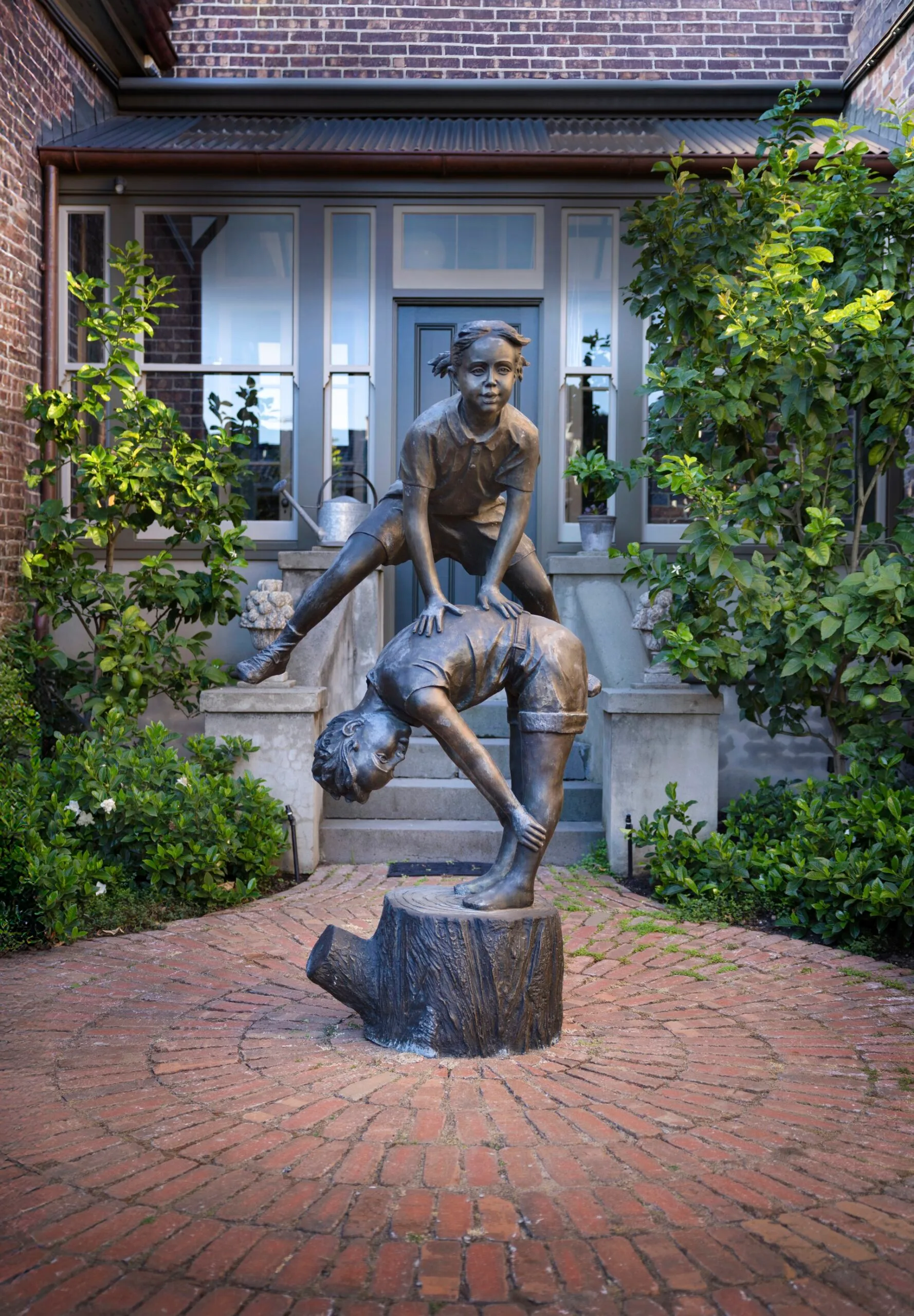 Bronze statue of two children playing leapfrog on a brick pathway in front of a brick house with greenery around.