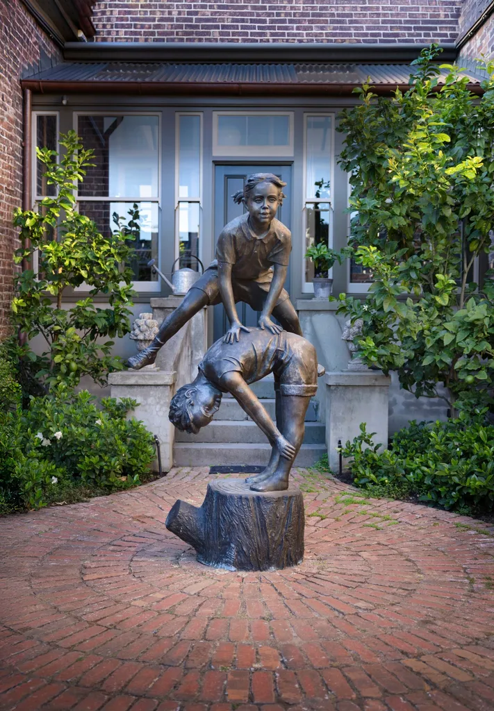 Bronze statue of two children playing leapfrog on a brick pathway in front of a brick house with greenery around.