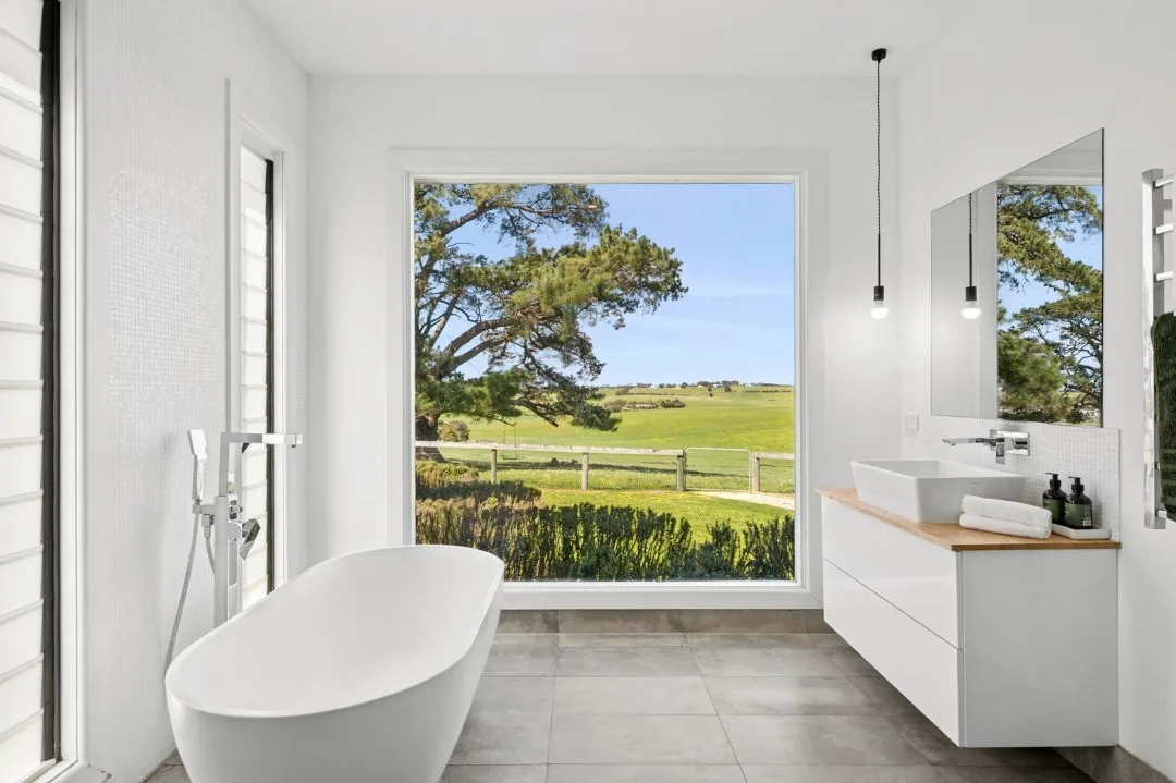 White bathroom featuring freestanding bathtub with picture window showing views of a farm and rolling hills.