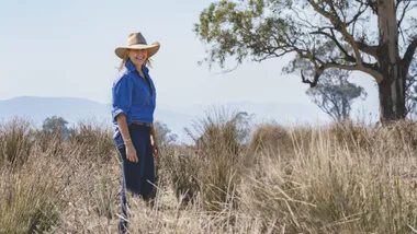 Woman in blue shirt and cowboy hat standing in grassy field, with trees and mountains in the background.