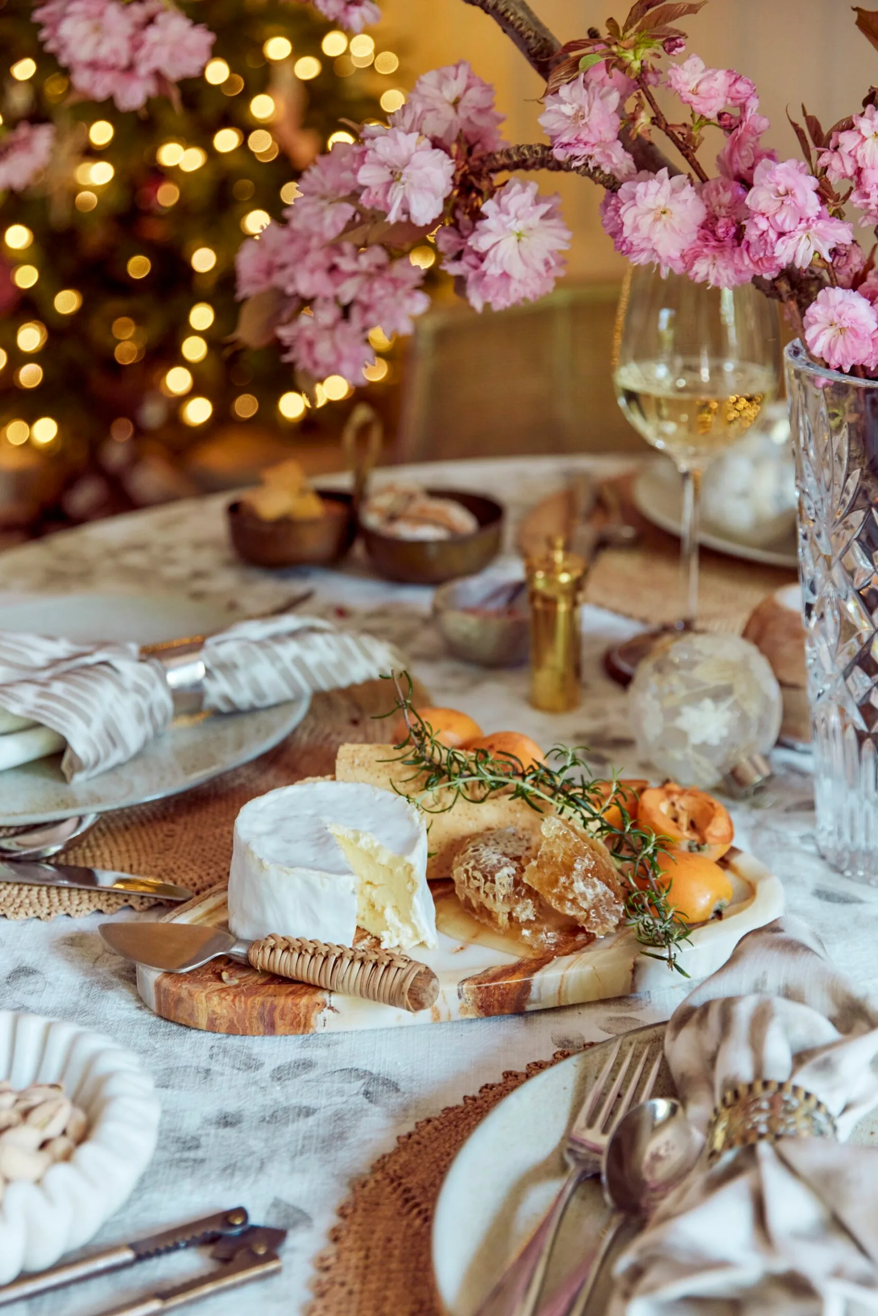A table with neutral linens, plates and gold napkin rings. Glasses are filled with white wine and there's a marble cheeseboard which has a wheel of soft cheese and honey on it. A crystal vase has a plant with pink flowers in it.