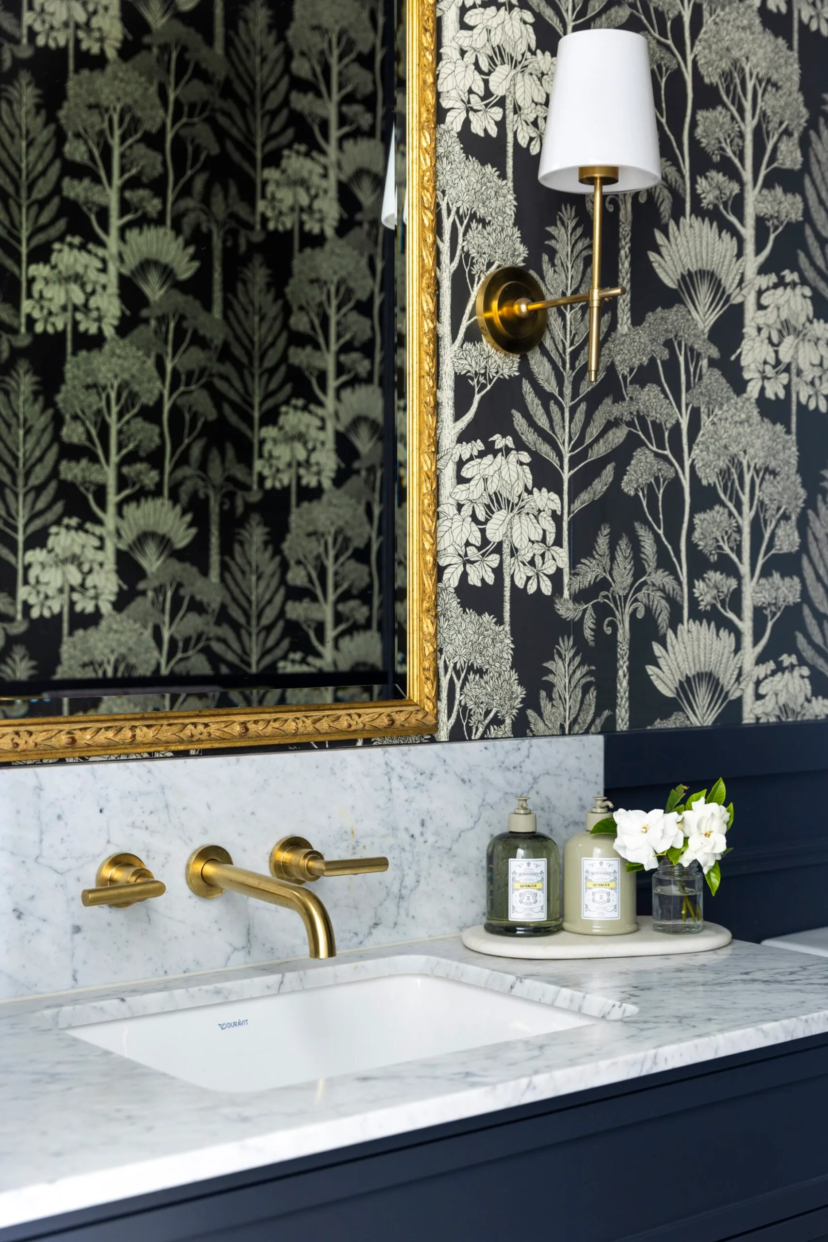 A powder room with a gold framed mirror, wall lights, marble benchtop and splashback and brass tapware.