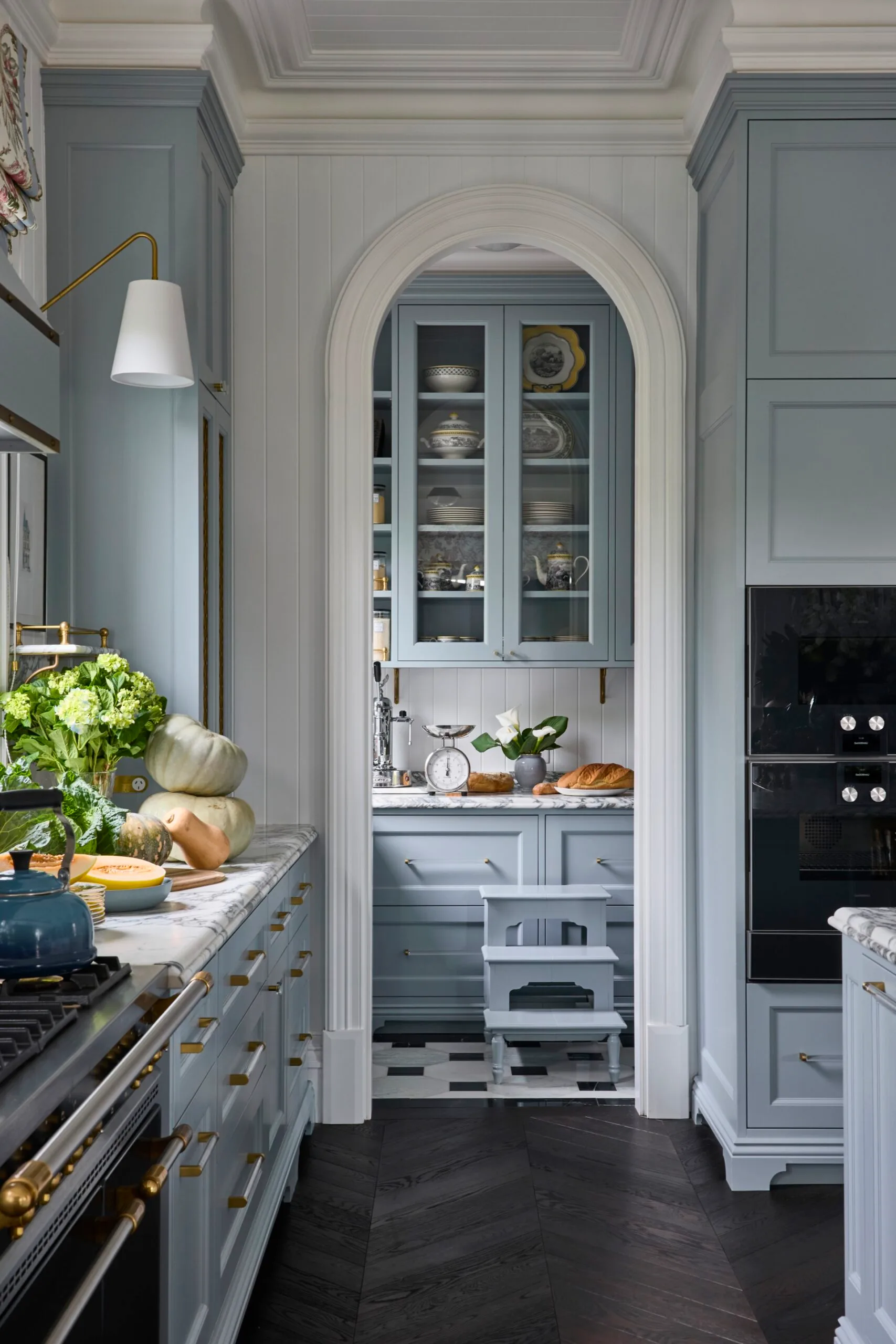 A kitchen with blue cabinetry, dramatically veined marble benchtops and splashbacks, brass accents and VJ panelled walls and ceiling. An arch leads to a butler's pantry with matching finishes.