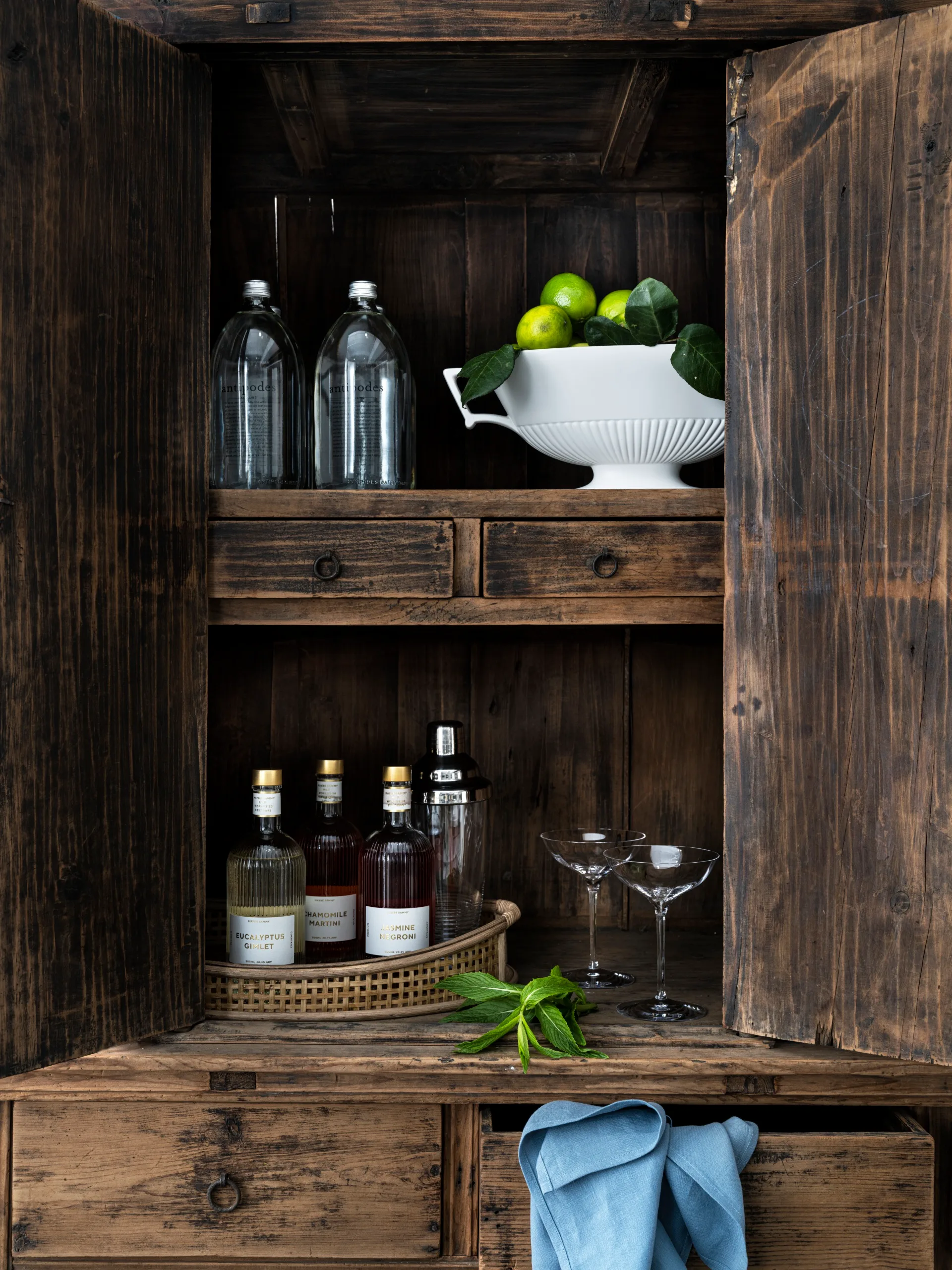 A wooden cupboard that looks rustic. THe doors of the cupboard are open, revealing a rattan tray filled with bottles of spirits and cocktail mixes, as well as two crystal coupes and a white bowl filled with limes. A blue tablecloth is folded over a drawer that is ajar, underneath the open cupboard doors.