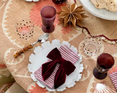 A table with light brown table cloth, white plates with straiped red and white napkins on top and red velvet bows on top of that. The glassware is a deep red, with matching baubles nearby. A pavlova topped with red grapes is in the centre of the table.