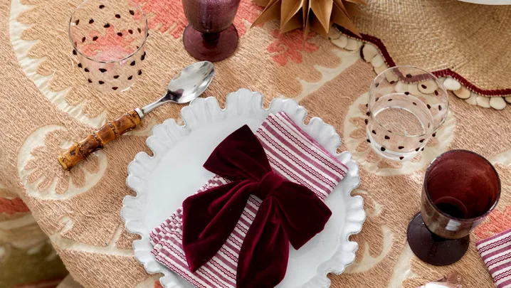 A table with light brown table cloth, white plates with straiped red and white napkins on top and red velvet bows on top of that. The glassware is a deep red, with matching baubles nearby. A pavlova topped with red grapes is in the centre of the table.
