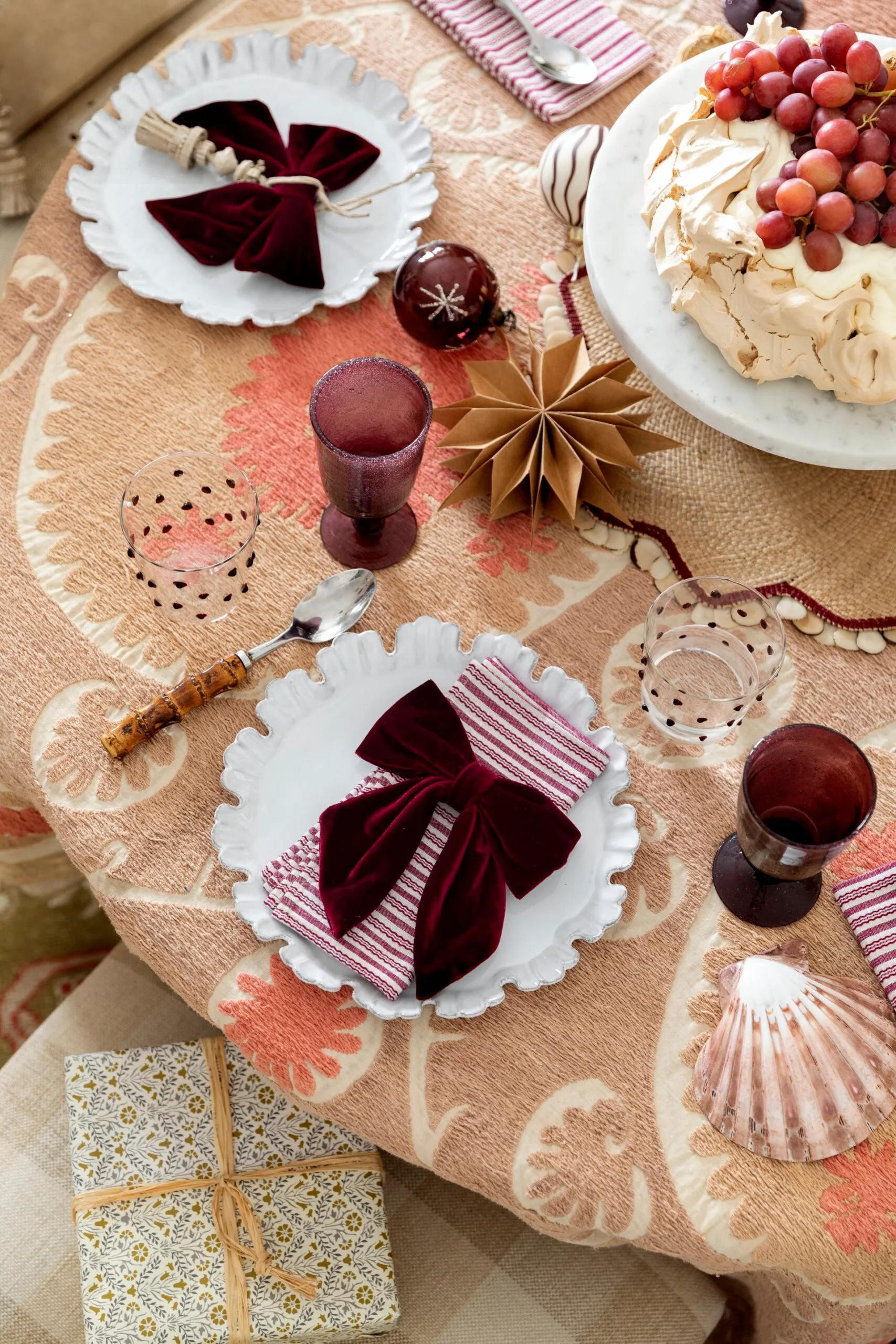 A table with light brown table cloth, white plates with straiped red and white napkins on top and red velvet bows on top of that. The glassware is a deep red, with matching baubles nearby. A pavlova topped with red grapes is in the centre of the table.