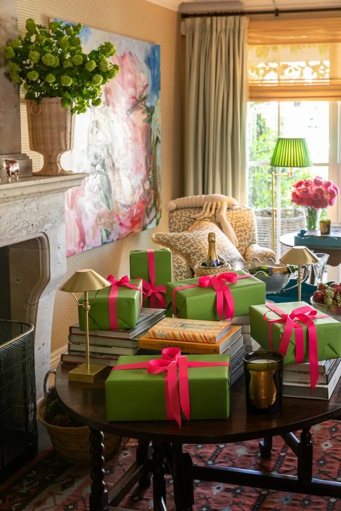 A living room which has a fireplace, a large artwork on the wall, and a round dark timber table. The table has hard cover books stacked on top, with lots of presents on top of that. The presents are wrapped in green wrapping paper with bright pink ribbons tied in bows.