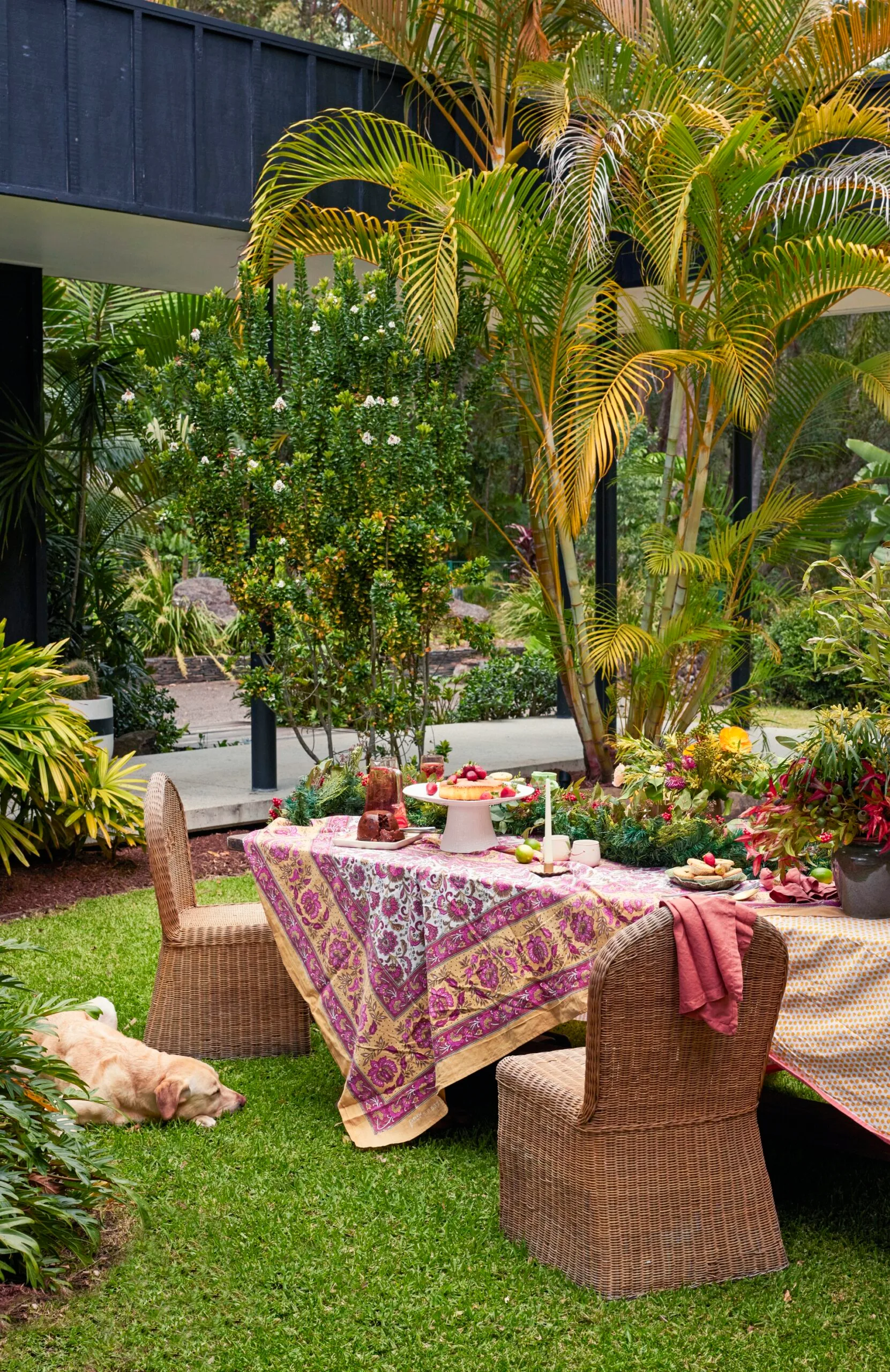 An outdoor dining table with woven dining chairs, pink and yellow tablecloths, and abundant greenery lining the middle of the table. A dog lies beside the table on the lawn.