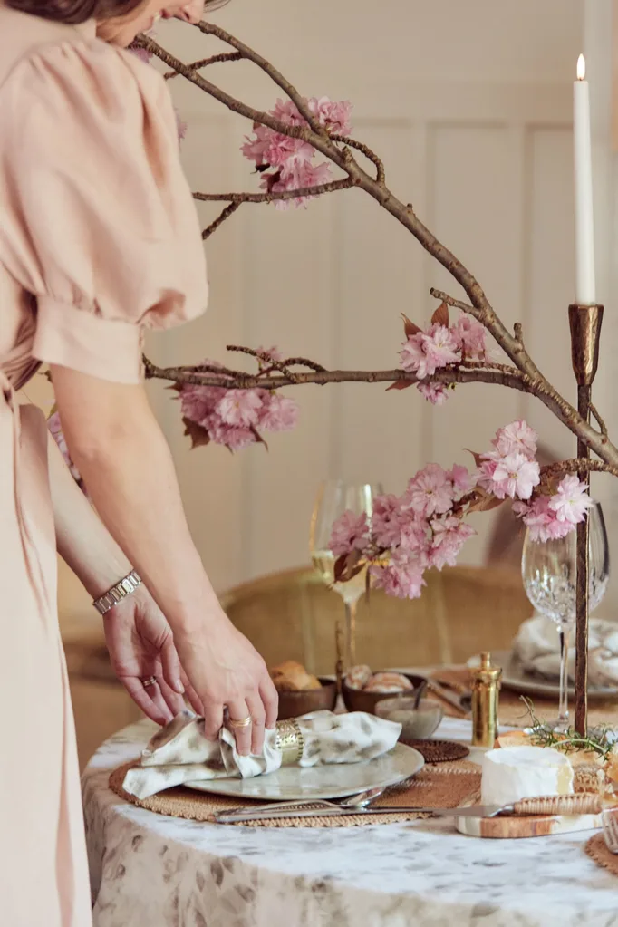 Designer and stylist Kate Nixon is half in the frame, wearing a light, dusty pink dress. She folds a napkin on top of a plate. The table is decorated with neutral linens, gold napkin rings, tall brass candlesticks with a tapered candle lit. Wine glasses are filled with white wine and a plant that has long branches, decorated with pink flowers, is the centrepiece.