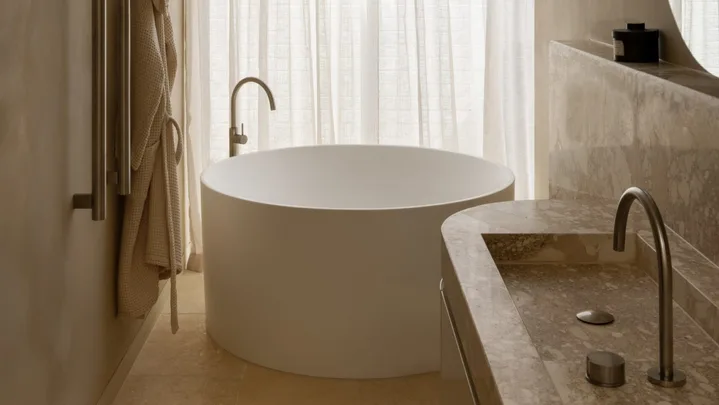 Modern bathroom with a round bathtub, minimalist fixtures, towel rack, and marble countertop beside sheer curtains.