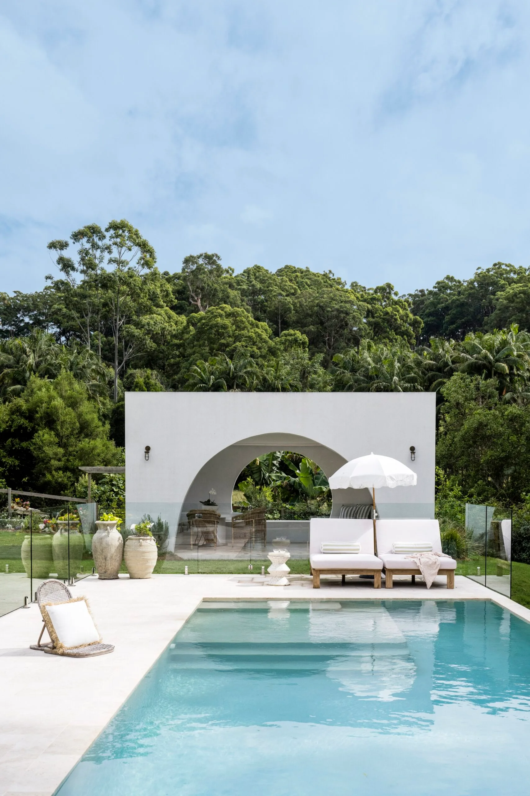 Pool and cabana with loungers and an umbrella among lush landscaping.