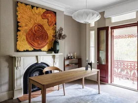 Office with wooden desk, chair, ornate fireplace, abstract painting, and open double doors to a balcony.