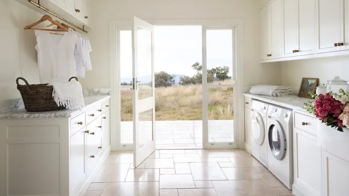 Laundry room with white cabinets (overhead and underbench), marble countertops, two washing machines, and a door opening to a grassy, rural Australian field outside. The floor is tiled and the joinery has brass knobs and a shaker profile.