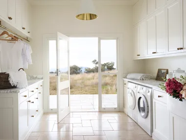 Laundry room with white cabinets (overhead and underbench), marble countertops, two washing machines, and a door opening to a grassy, rural Australian field outside. The floor is tiled and the joinery has brass knobs and a shaker profile.