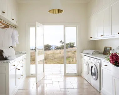 Laundry room with white cabinets (overhead and underbench), marble countertops, two washing machines, and a door opening to a grassy, rural Australian field outside. The floor is tiled and the joinery has brass knobs and a shaker profile.