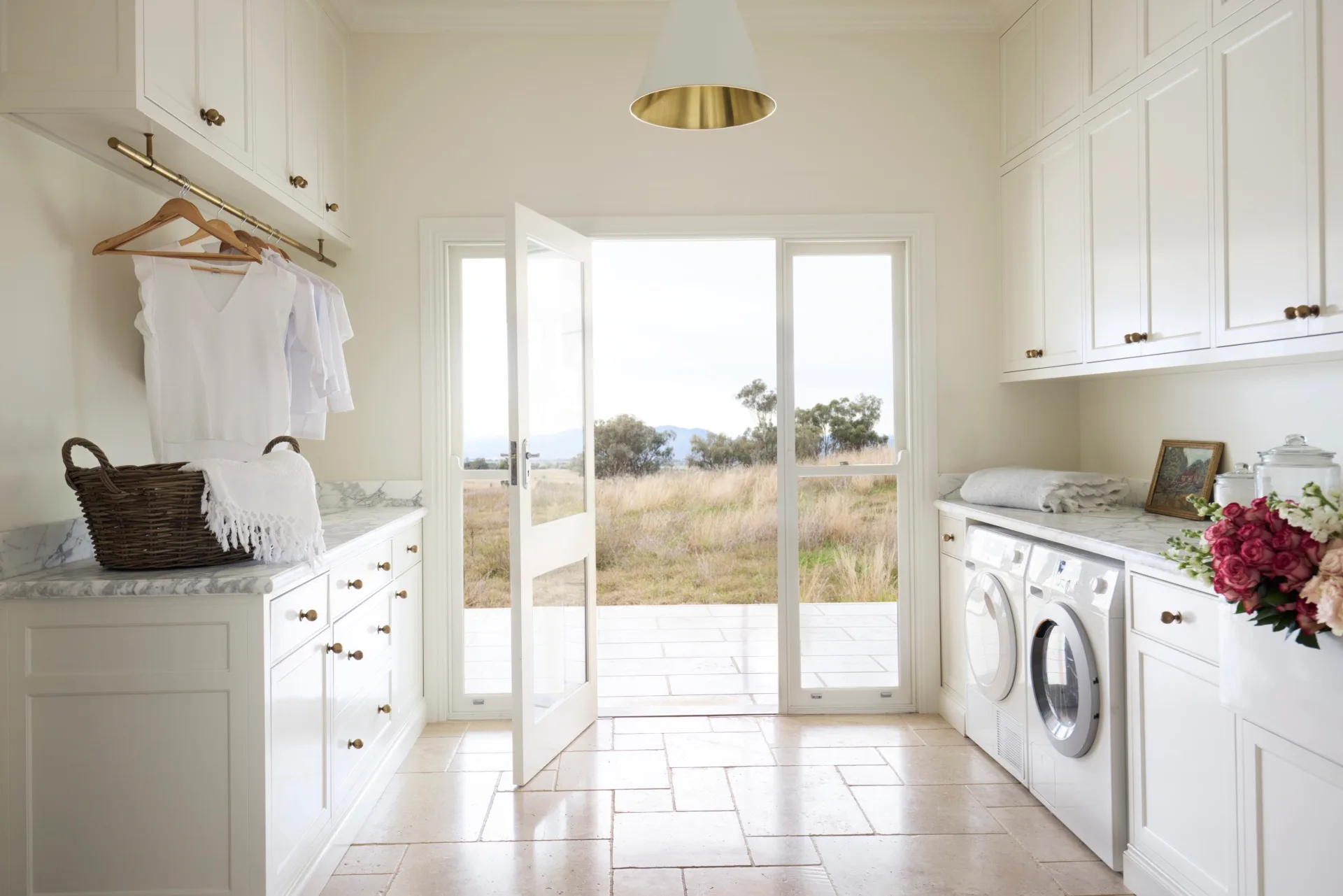 Laundry room with white cabinets (overhead and underbench), marble countertops, two washing machines, and a door opening to a grassy, rural Australian field outside. The floor is tiled and the joinery has brass knobs and a shaker profile.