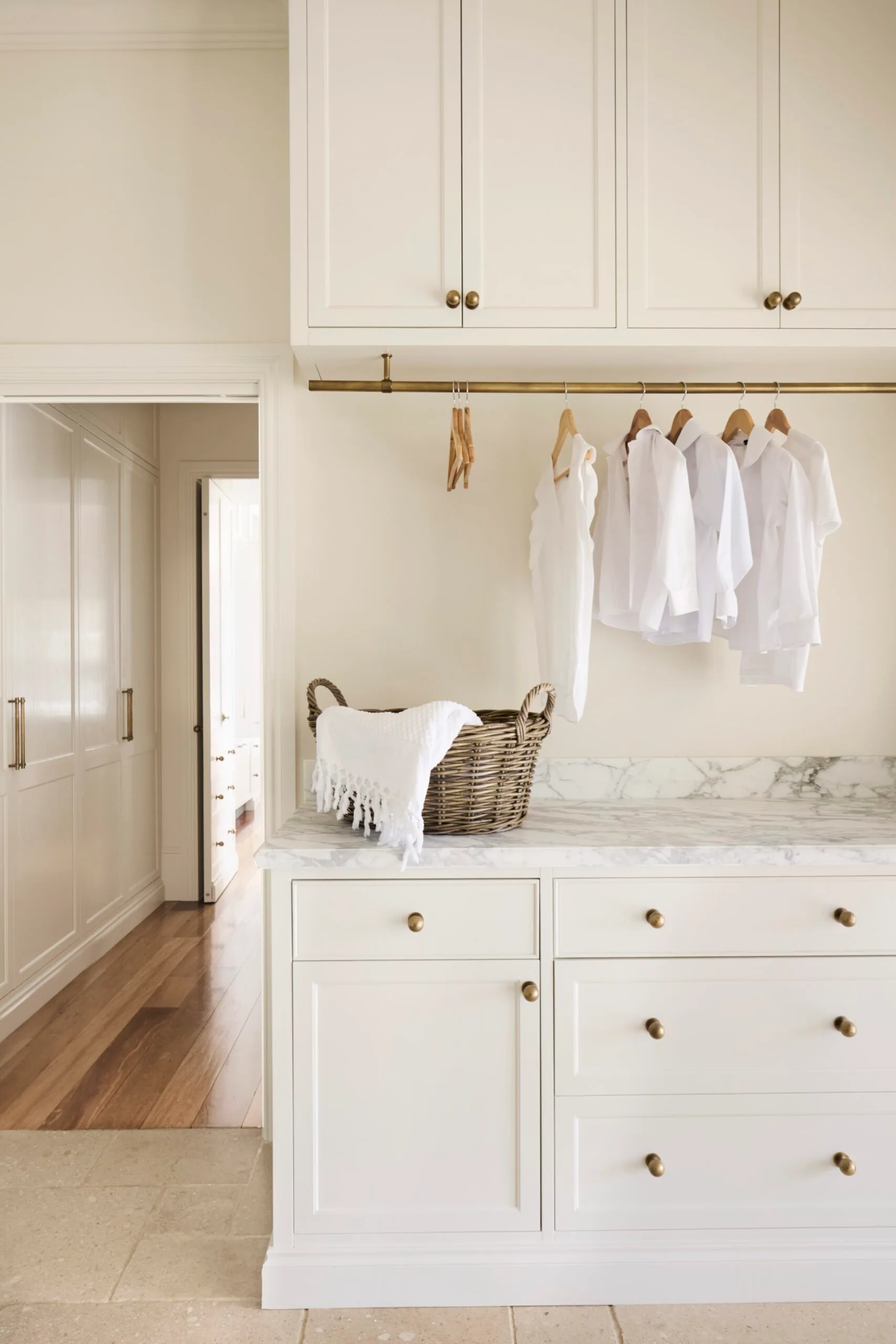 Laundry room with white cabinets (overhead and underbench), marble countertops, a wicker basket on the counter and a brass hanging rod where clothing can hang to air dry. The floor is tiled and the joinery has brass knobs and a shaker profile. An open door to the left leads to a hallway with wooden floorboards.