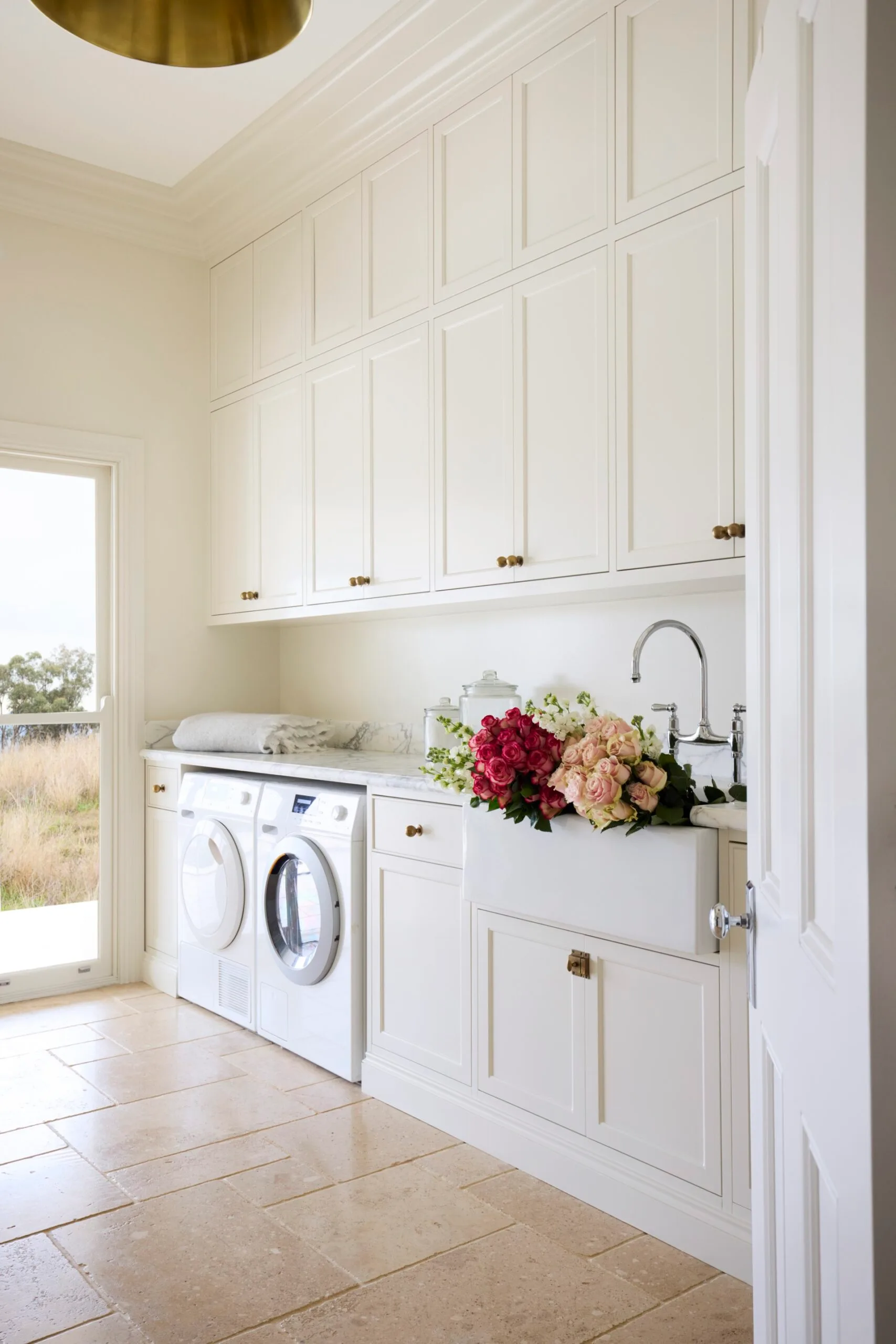 Laundry room with white cabinets (overhead and underbench), marble countertops, two washing machines, and a door opening to a grassy, rural Australian field outside. The floor is tiled and the joinery has brass knobs and a shaker profile.