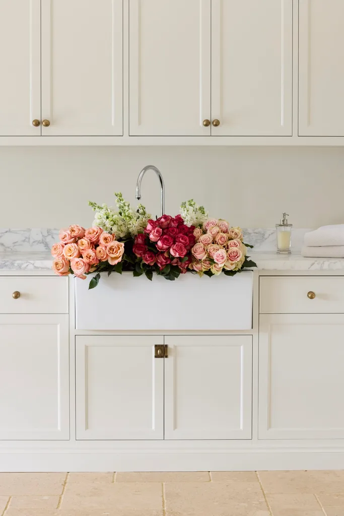 Laundry room with white cabinets (overhead and underbench), marble countertops and a large white ceramic farmhouse sink. Pink and red roses are placed overflowing in the sink, with white flowers as well. The floor is tiled and the joinery has brass knobs and a shaker profile.