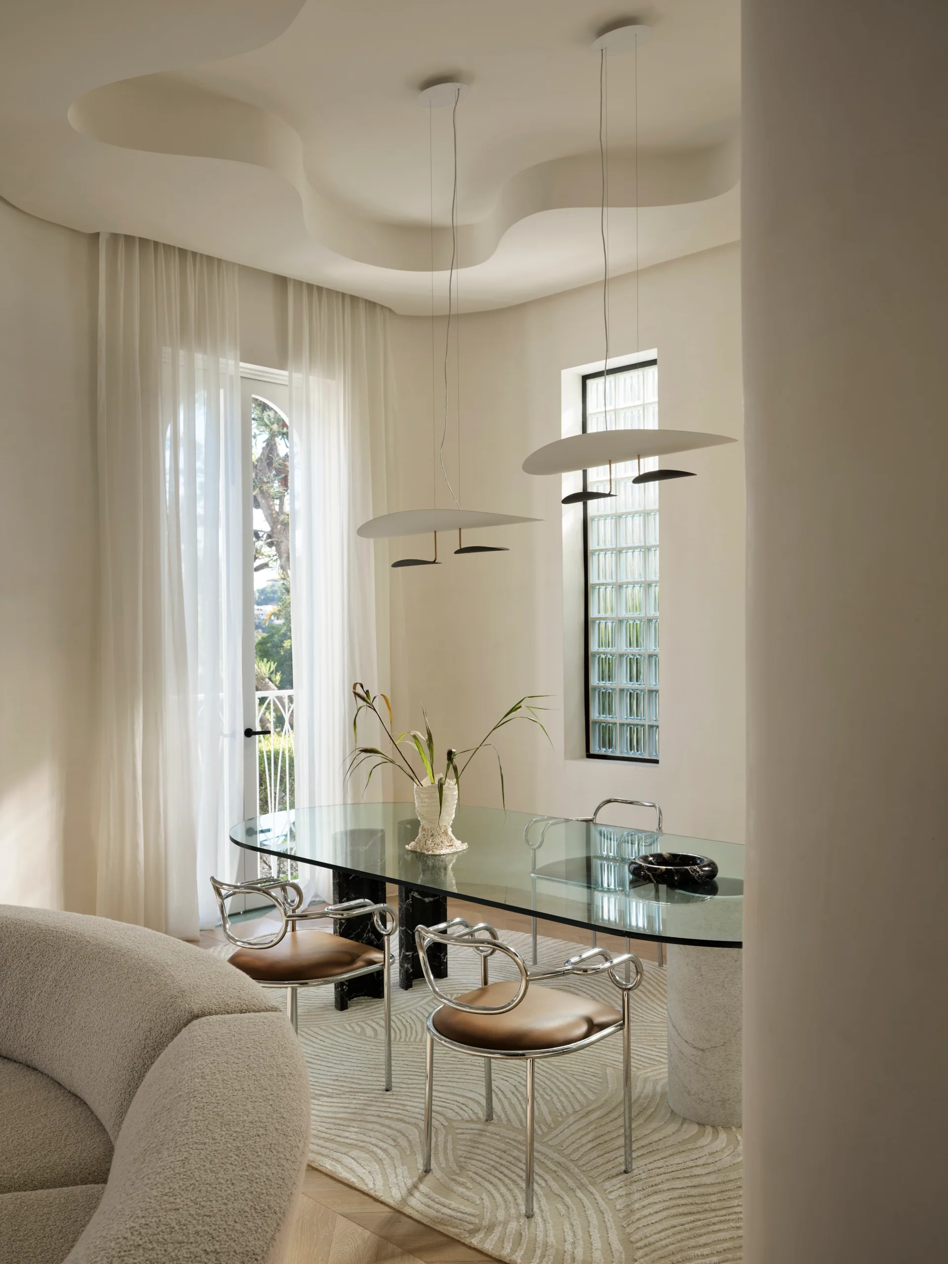 Modern dining room with glass table, sculptural chairs, a ceiling with a raised section in the middle, which has a wavy shape. A tall window has sheer curtains and hanging from the tall ceiling are two modern light pendants.