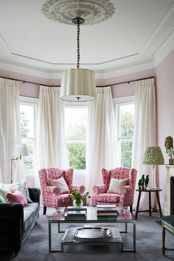 Elegant living room with pink walls, patterned armchairs, large windows with white curtains, and a decorative pendant lighting hanging from a ceiling rose.
