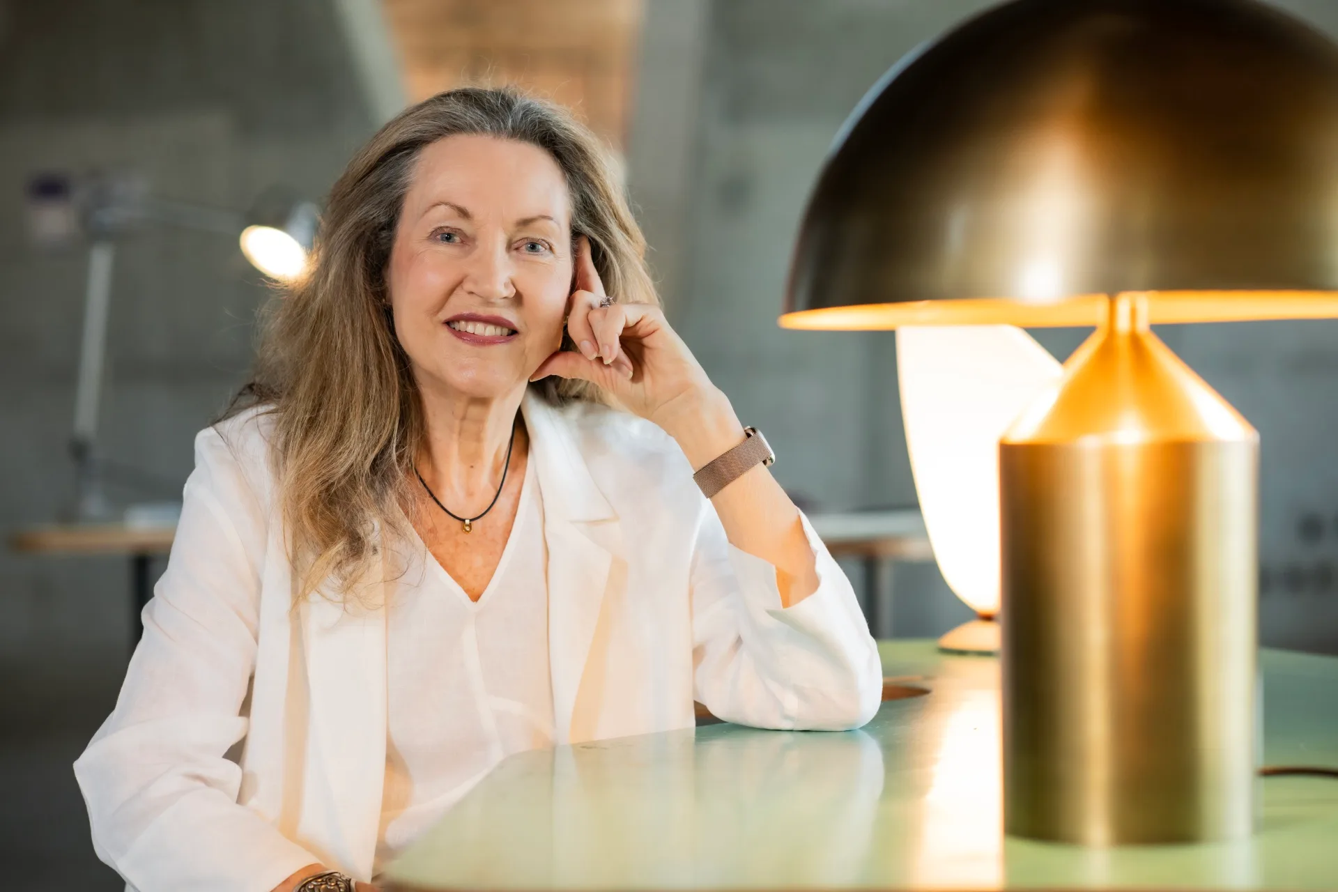A photo of a woman wearing a white blouse and matching blazer. She is sitting at a table with a table lamp in the foreground and another behind her. The one in the foreground has a mushroom shape and is made of brass.
