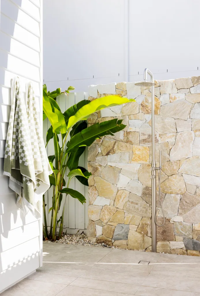Outdoor shower against a stone feature wall outside a white weatherboard house with green checkerboard towels and plants.