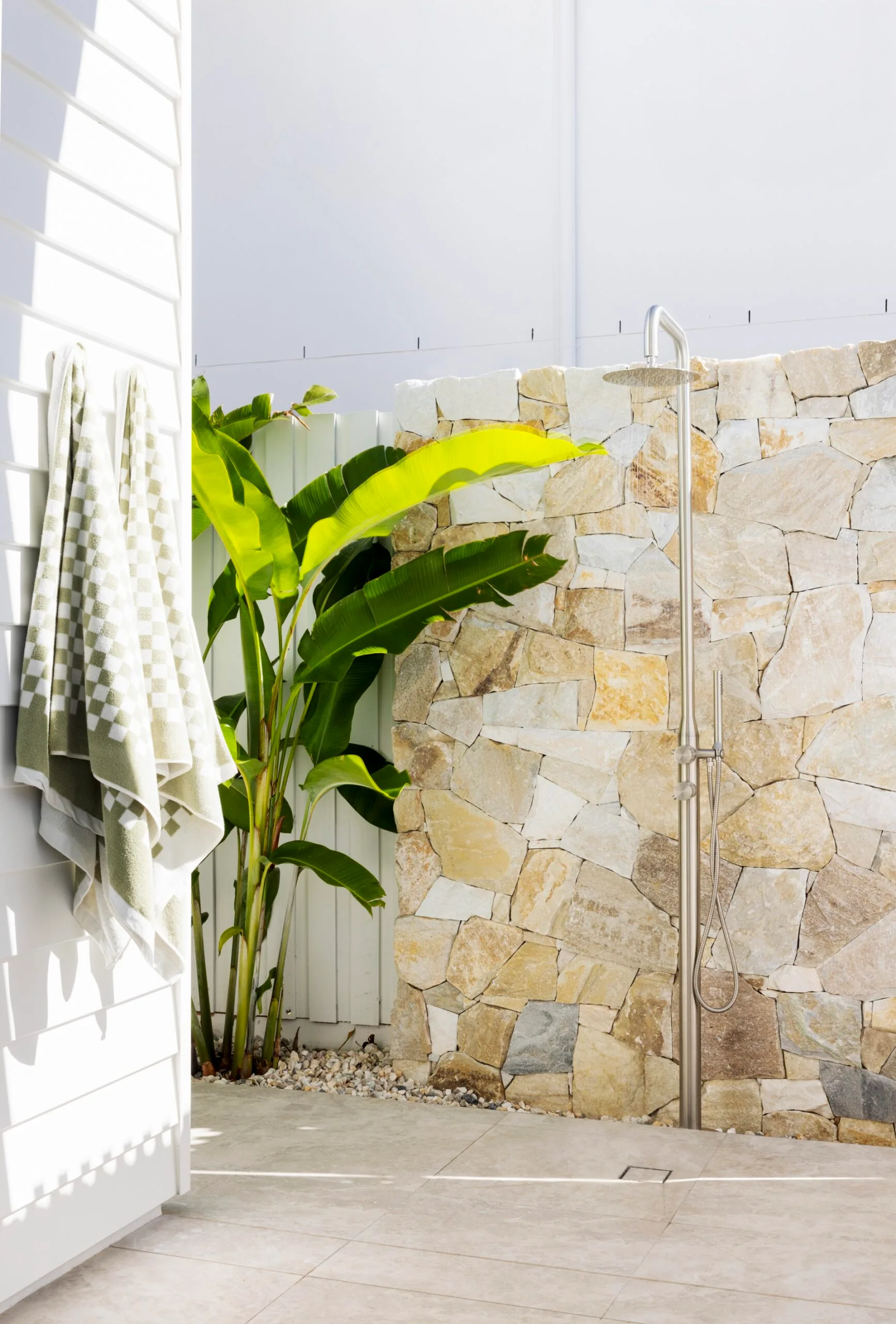 Outdoor shower against a stone feature wall outside a white weatherboard house with green checkerboard towels and plants.