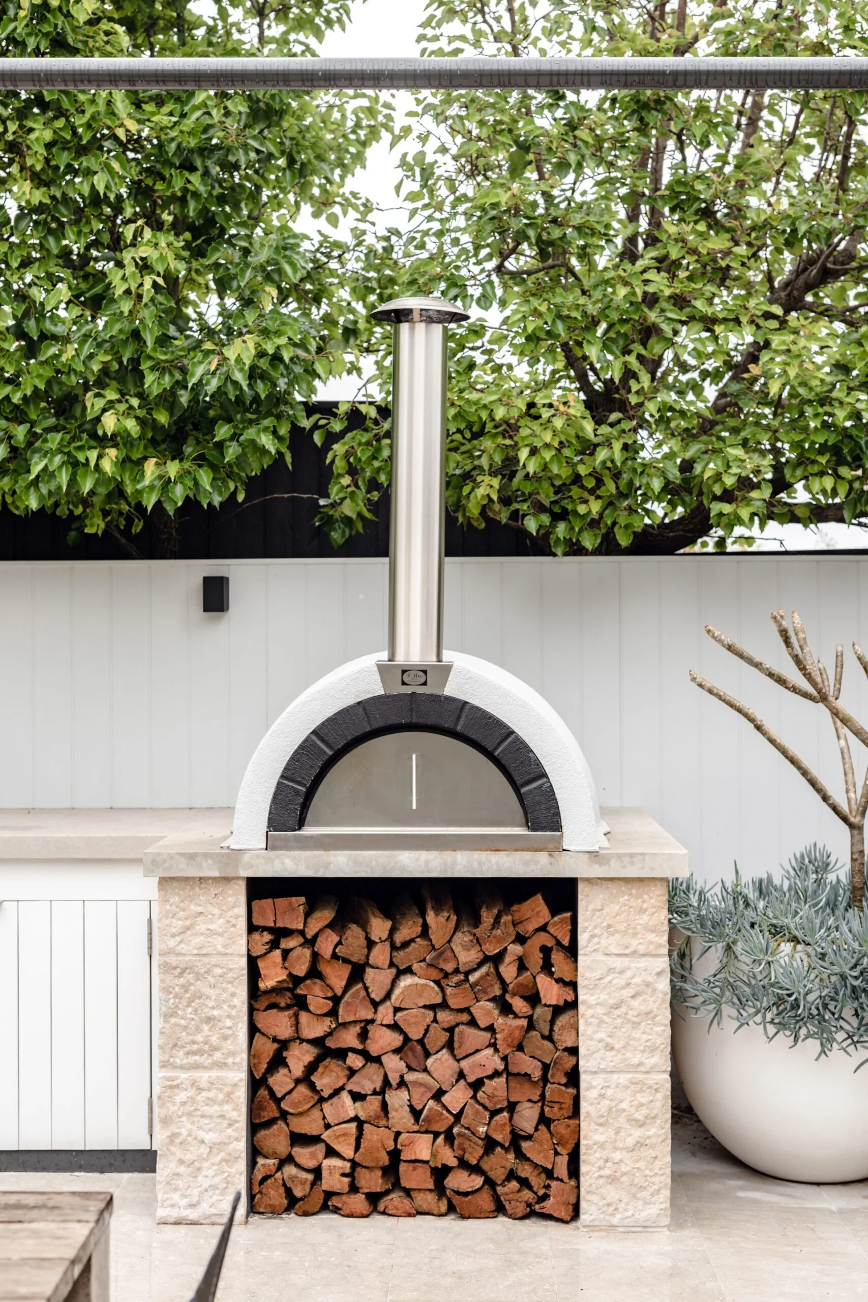 A wood-fired pizza oven in an outdoor kitchen, with trees growing above a fence in the background and a large potted plant beside the pizza oven.