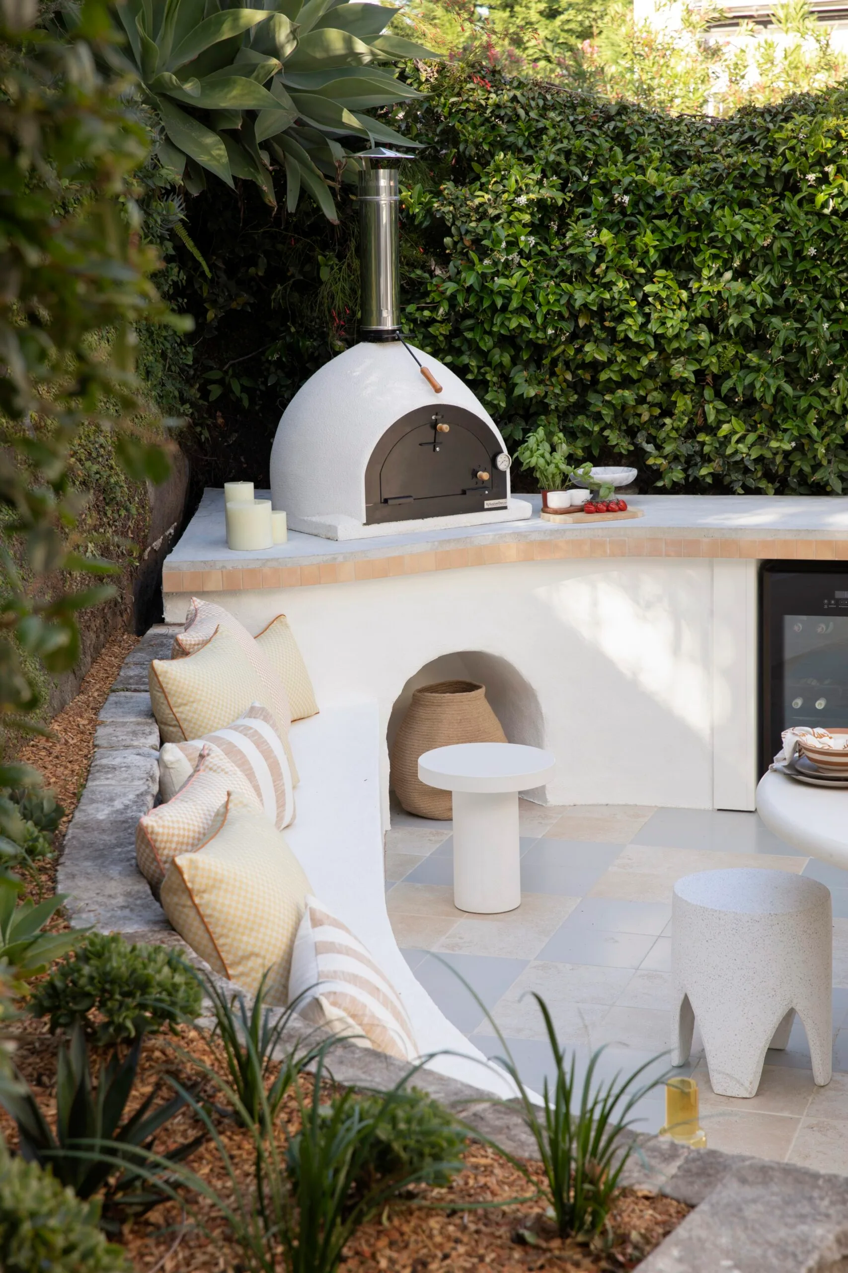 An outdoor kitchen. A curved built-in bench has cushions on it, extending around from the kitchen. A green hedge grows around, providing privacy.