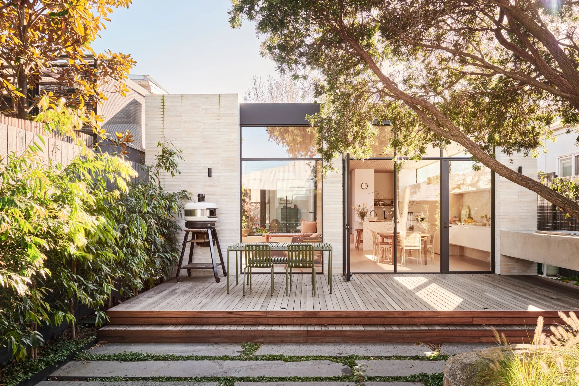An outdoor kitchen and dining area with a freestanding pizza oven to the left. The back of the house has glass doors and an indoor kitchen is visible through these. Lush greenery is visible, growing beyond the deck where the outdoor kitchen and dining area is.