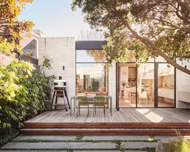 An outdoor kitchen and dining area with a freestanding pizza oven to the left. The back of the house has glass doors and an indoor kitchen is visible through these. Lush greenery is visible, growing beyond the deck where the outdoor kitchen and dining area is.