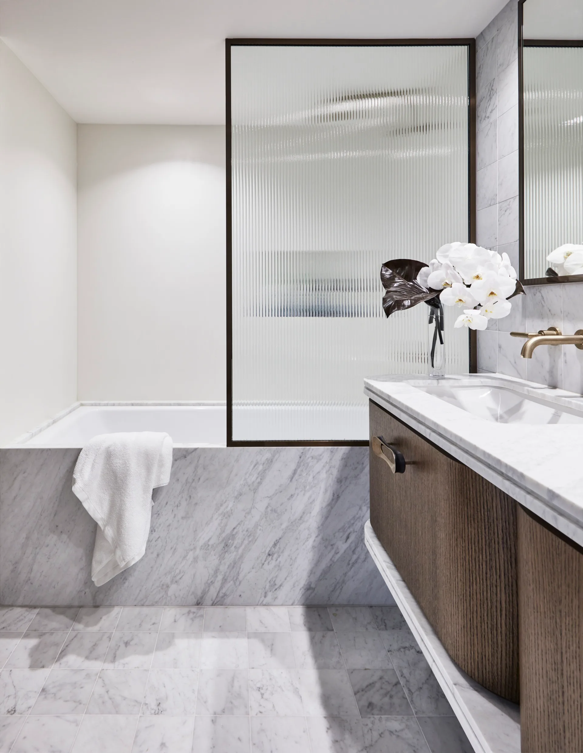 Ensuite bathroom with tub, wooden vanity, white towel, and vase of white flowers. The bathtub is built in and the bath fascia is marble, as is the flooring and the vanity benchtop. A shower screen is framed in black and the timber vanity has brass handles.
