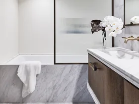 Ensuite bathroom with tub, wooden vanity, white towel, and vase of white flowers. The bathtub is built in and the bath fascia is marble, as is the flooring and the vanity benchtop. A shower screen is framed in black and the timber vanity has brass handles.