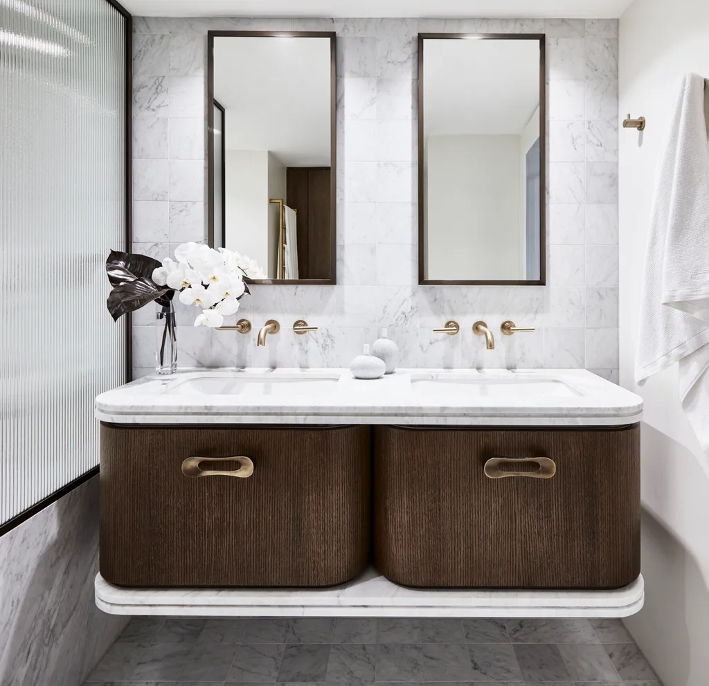 An ensuite bathroom with marble wall tiles, floor tiles and bath fascia. A glass screen blocks the view of the shower to the left. The timber vanity has a curved edge and marble benchtop. THe hardware is brass and two rectangle mirrors are positioned above twin basins set into the marble benchtops.